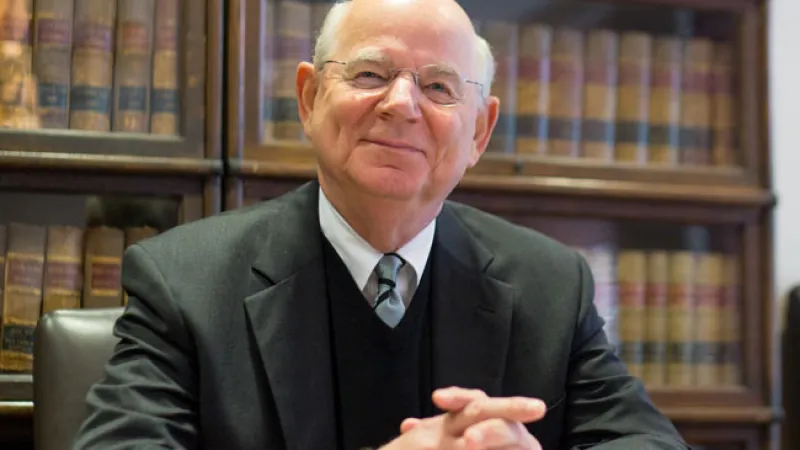 Elderly man in a suit sits at a desk with law books behind him, smiling slightly.