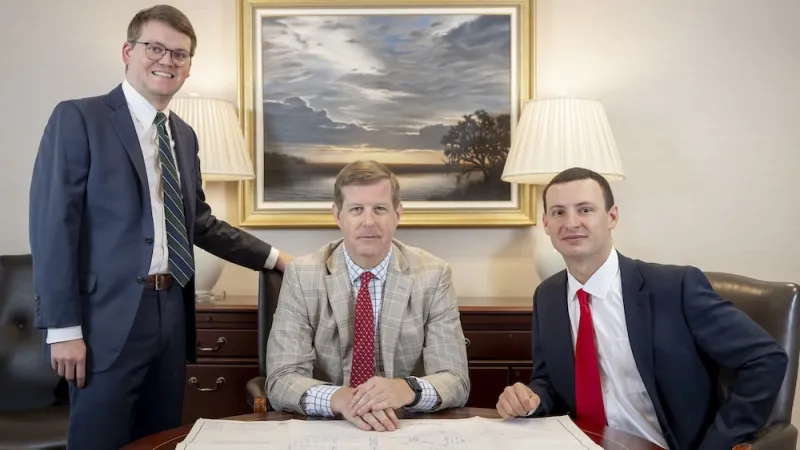 Three men in suits seated at a desk with a framed painting behind them.