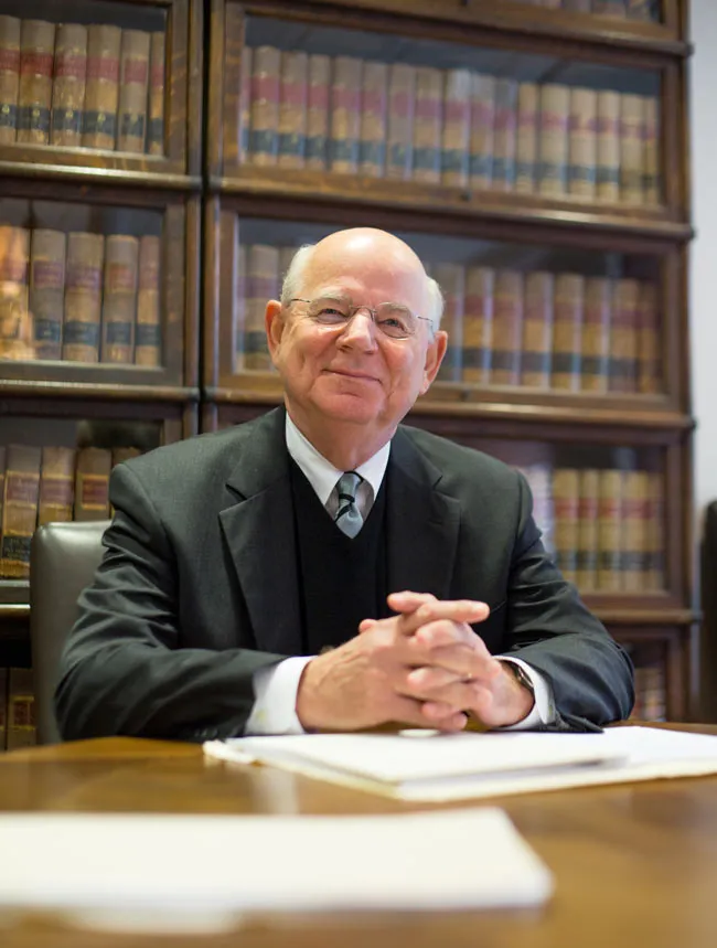 Elderly man in a suit sits at a desk with law books behind him, smiling slightly.