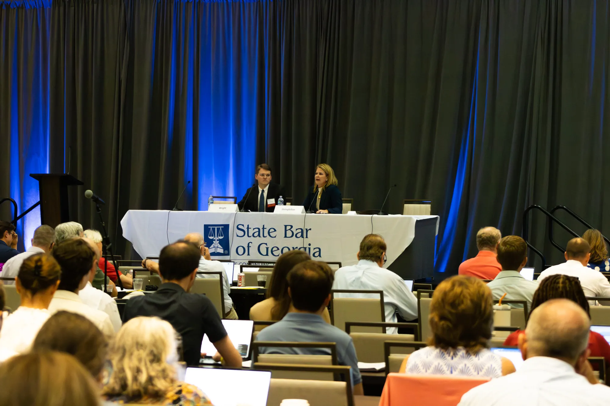 Audience listening to speakers at a conference with blue-lit curtains.